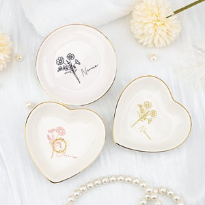 Könnte beinhalten: Drei weiße Keramik-Ringschalen mit goldenen Rändern. Eine runde Schale und zwei herzförmige Schalen werden gezeigt. Jede Schale hat ein Blumendesign und einen Namen. Eine herzförmige Schale enthält einen goldenen Ring. Die Schalen stehen auf einer weißen Oberfläche.