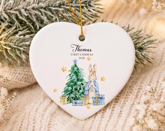 May include: A white heart-shaped ceramic ornament with a gold hanging string. The design features a watercolor illustration of a rabbit, a Christmas tree, and gifts, with the text "Thomas FIRST CHRMAS 2026" printed in black ink above the graphic.