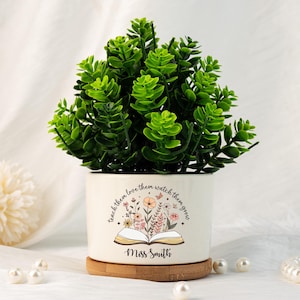 May include: A small, round, white ceramic planter with a green plant. The planter has a floral design and the text "teach them love them watch them grow" and "Miss Smith". The planter sits on a round, wooden coaster.