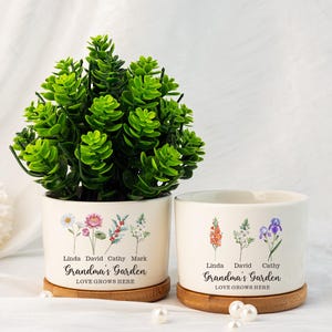 Könnte beinhalten: Zwei weiße Keramik-Blumentöpfe mit floralem Design und dem Text "Grandma's Garden, Love Grows Here". Die Töpfe stehen auf einem Holzsockel und tragen die Namen Linda, David, Cathy und Mark.