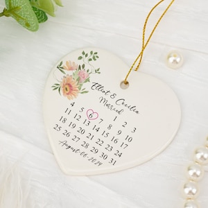 Puede incluir: Un adorno de cerámica blanca en forma de corazón con una ilustración floral, el texto Elliot & Cecilia Married y un calendario de agosto de 2024 con un corazón rosa rodeando el número 6. Incluye un cordón dorado para colgar el adorno.