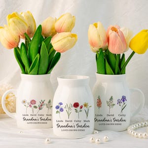 Op de afbeelding: Drie witte keramische vazen met handvatten, elk versierd met een bloemenontwerp en de tekst "Grandma's Garden, Love Grows Here". De vazen zijn gevuld met gele en roze tulpen.