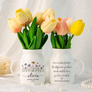 Puede incluir: Dos jarrones de cerámica blanca con asas, cada uno con un ramo de tulipanes en tonos amarillos, rosas y blancos. Un jarrón está personalizado con el nombre "Olivia" y el texto "Officially Retired 2025". El otro jarrón tiene el texto "Nunca subestimes la diferencia que has hecho y las vidas que has tocado."