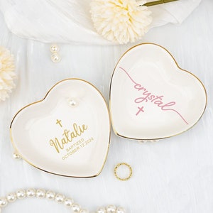 Könnte beinhalten: Zwei weiße herzförmige Keramik-Schmuckschalen mit goldenen Rändern. Eine Schale trägt die Aufschrift "Natalie Baptized October 12, 2024" in goldenen Buchstaben mit einem goldenen Kreuz. Die andere Schale trägt die Aufschrift "crystal" in rosa Buchstaben mit einem goldenen Kreuz.