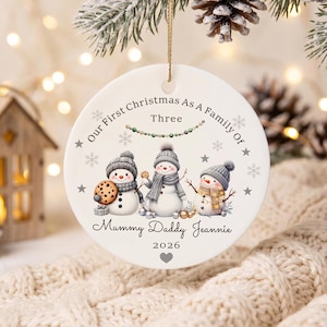 May include: A round white ceramic Christmas ornament featuring three illustrated snowmen wearing grey hats and scarves. The text reads Our First Christmas As A Family Of Three, Mummy Daddy Jeannie, and 2026, accented with grey stars and snowflakes.