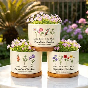 May include: Three cream-colored ceramic planters with floral designs and the text "Grandma's Garden." Each planter holds pink flowering plants and sits on a round wooden base. The text "Love Grows Here" is also printed on the planters.