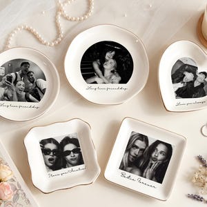 May include: Five white ceramic trinket dishes with gold-trimmed edges, each featuring a black and white photograph and printed text. The dishes are round, heart-shaped, and square, displaying various personal photos and phrases like Long live friendship.
