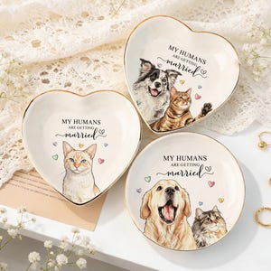 Peut inclure: Trois coupelles en céramique avec des bords dorés. Deux sont en forme de cœur et une est circulaire. Chacune présente une illustration colorée de chien et de chat, des petits cœurs et le texte imprimé : MY HUMANS ARE GETTING married.
