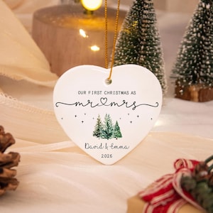 May include: A white heart-shaped ceramic ornament featuring the text "OUR FIRST CHRISTMAS AS mr & mrs" with a small heart icon, a watercolor illustration of three pine trees, and the names "David & Emma" with the year "2026" printed below.
