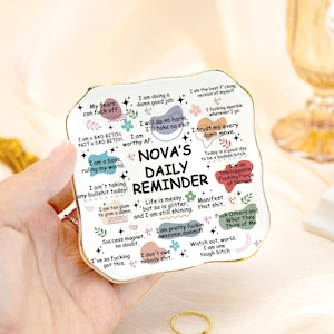 Puede incluir: Un plato recordatorio diario de cerámica blanca con borde dorado, con varias frases empoderadoras y el texto "NOVA'S DAILY REMINDER". El plato tiene forma rectangular con bordes redondeados y detalles florales.