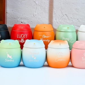 May include: A collection of ceramic pet urns in various colors, shaped like cat faces. Each urn has a different name and design, including "Jasper," "Lucky," "Love Honey," "Pony," "Simba," "Peanut," "Cooper," and "Teddy."