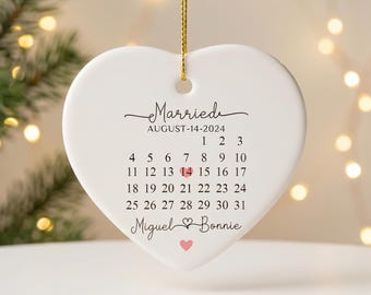 May include: A white heart-shaped ceramic ornament hanging from a gold string. It features the text Married, August-14-2024, a calendar for August with the 14th highlighted by a pink heart, and the names Miguel and Bonnie with a small heart.