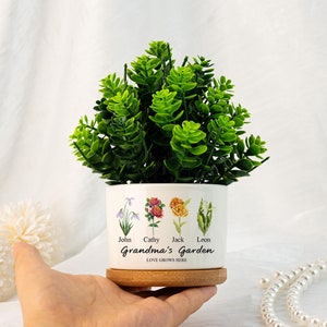 May include: White ceramic planter with a wooden base, featuring a green artificial plant. The planter has a floral design with four flowers and the names John, Cathy, Jack, and Leon. The text "Grandma's Garden" and "Love Grows Here" are printed on the planter.
