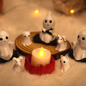 Peut inclure: Une composition sur le thème d'Halloween avec des figurines de fantômes blancs, des figurines d'animaux et une bougie allumée. Les figurines sont disposées sur une base en bois et une surface blanche et moelleuse, avec une citrouille en arrière-plan.