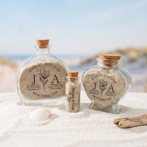 Puede incluir: Tres botellas de vidrio llenas de arena, cada una con un tapón de corcho. Las botellas tienen diferentes formas: redonda, corazón y vial pequeño. Las botellas tienen texto personalizado: nombres, una fecha y ubicación. Las botellas están en una playa arenosa.