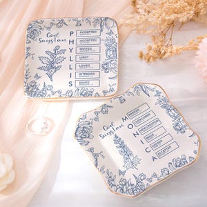 Puede incluir: Dos platos cuadrados de cerámica blanquecina con ribete dorado, con diseños florales azules y texto. Un plato dice "God Sayslam" y "PHYLLIS" con palabras relacionadas. El otro dice "God Sayslam" y "MONICA".