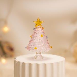 Op de afbeelding: Een klein, conisch kerstboomfiguur van glas in een zachtroze tint met een geribbelde textuur. De boom is bekroond met een heldergele glazen ster en versierd met kleine, verspreide gele en blauwe glazen accenten, alles op een voet van helder glas.