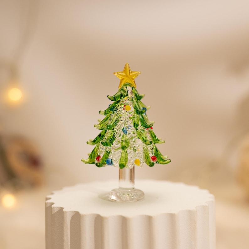 Op de afbeelding: Een miniatuur kerstboomfiguur van glas met een heldere, getextureerde stam en voet. De boom heeft groene glazen takken versierd met kleine, kleurrijke glazen bollen en is bekroond met een heldere gele glazen ster als piek.