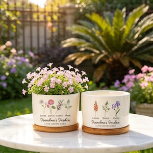 K&ouml;nnte beinhalten: Zwei cremefarbene Keramik-Blument&ouml;pfe mit floralen Designs und dem Text "Grandma's Garden". Ein Topf enth&auml;lt rosa bl&uuml;hende Pflanzen. Beide stehen auf Bambus-Untersetzern. Der Text "LOVE GROWS HERE" ist ebenfalls aufgedruckt.