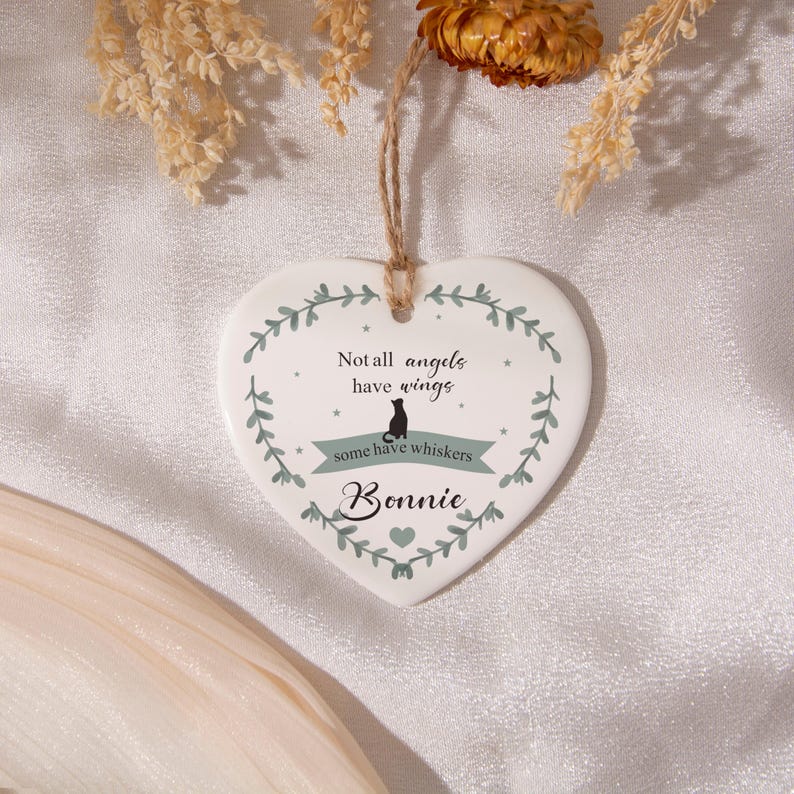 Puede incluir: Un adorno de cer&aacute;mica blanco en forma de coraz&oacute;n que cuelga de un cordel de yute. Presenta un dise&ntilde;o de corona de hojas verdes, la silueta de un gato negro y el texto: Not all angels have wings, some have whiskers, y el nombre Bonnie en cursiva.