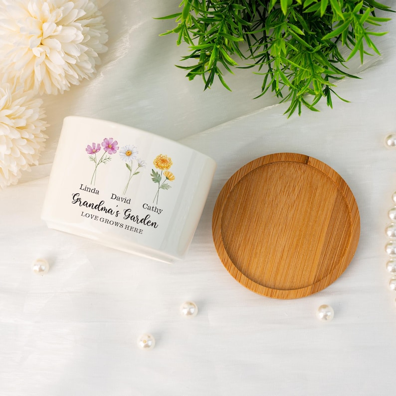 K&ouml;nnte beinhalten: Ein wei&szlig;er Keramik-Pflanztopf mit einer Holzuntertasse. Der Pflanztopf zeigt Blumenillustrationen und den Text "Grandma's Garden LOVE GROWS HERE" mit Namen. Das Design umfasst rosa, wei&szlig;e und gelbe Blumen.
