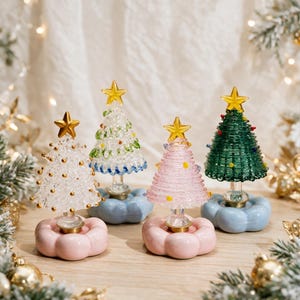 Op de afbeelding: Vier decoratieve kerstboombeeldjes van glas, elk bekroond met een gouden ster. De bomen hebben gevarieerde texturen en kleuren, waaronder transparant, wit, roze en groen, en zijn gemonteerd op afgeronde, wolkachtige voetstukken.