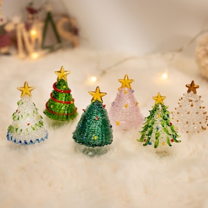 Op de afbeelding: Een collectie van zes miniatuur kerstbomen van glas in diverse kleuren, waaronder transparant, groen en roze. Elke boom is voorzien van een goudkleurige ster als piek en decoratieve glazen accenten, en staat als los ornament.