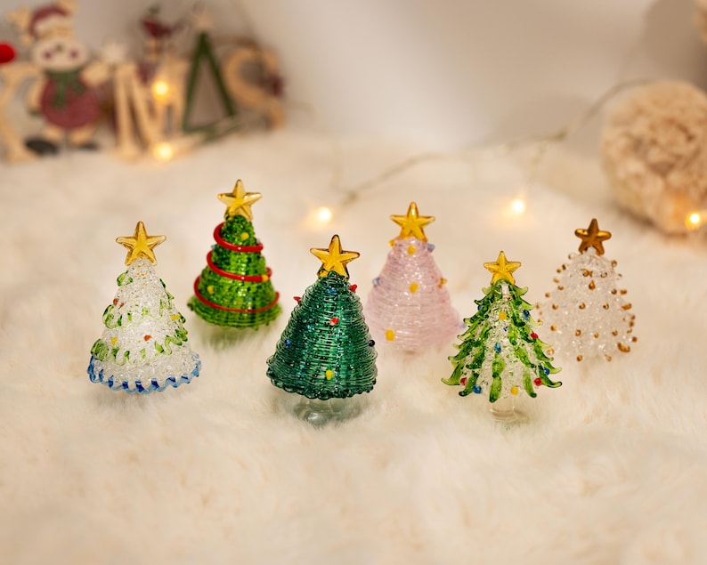 Op de afbeelding: Een collectie van zes miniatuur kerstbomen van glas in diverse kleuren, waaronder transparant, groen en roze. Elke boom is voorzien van een goudkleurige ster als piek en decoratieve glazen accenten, en staat als los ornament.