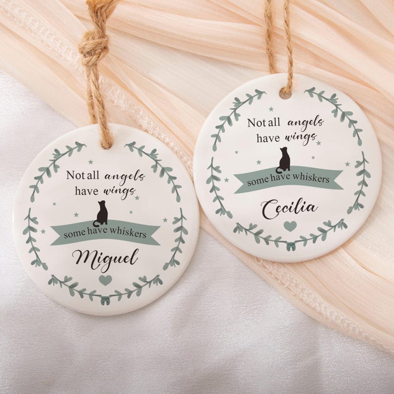 Puede incluir: Dos adornos redondos de cer&aacute;mica blanca con colgadores de cuerda de yute. Cada uno presenta una corona de hojas verdes, la silueta de un gato negro y el texto "Not all angels have wings, some have whiskers." Un adorno est&aacute; personalizado con "Miguel" y el otro con "Cecilia."