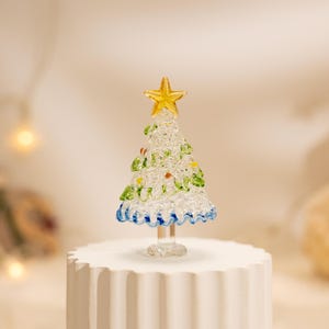 Op de afbeelding: Een kerstboomfiguur van helder glas met een getextureerd, kristallijn oppervlak met groene glazen accenten en kleine kleurrijke stippen. De boom is bekroond met een gele glazen ster en staat op een heldere glazen voet.