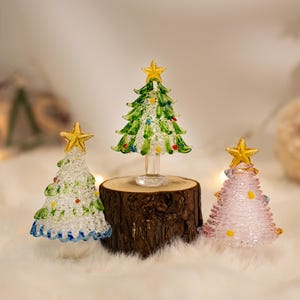 Op de afbeelding: Drie glazen kerstboombeeldjes met gouden sterren als piek. E&eacute;n boom is doorzichtig met groene en blauwe accenten, &eacute;&eacute;n is groen met veelkleurige stippen en &eacute;&eacute;n is lichtroze met gele stippen. De middelste boom staat op een kleine houten voet.