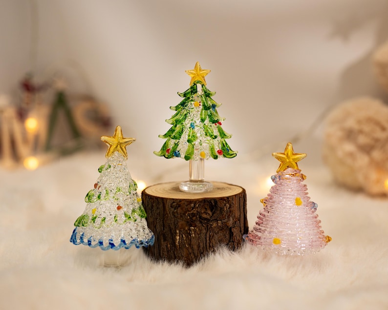 Op de afbeelding: Drie glazen kerstboombeeldjes met gouden sterren als piek. E&eacute;n boom is doorzichtig met groene en blauwe accenten, &eacute;&eacute;n is groen met veelkleurige stippen en &eacute;&eacute;n is lichtroze met gele stippen. De middelste boom staat op een kleine houten voet.