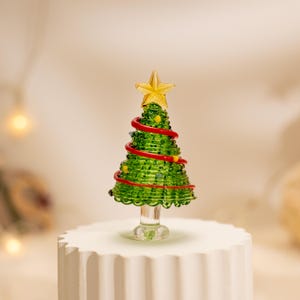 Op de afbeelding: Een miniatuur kerstboomfiguur van glas met een getextureerd groen conisch lichaam, een spiraalvormige rode glasslinger, kleine veelkleurige glaskralen als versiering en een doorzichtige ster van glas bovenop een glazen voetstuk.