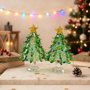Op de afbeelding: Twee glazen kerstboomfiguren met groene takken, heldere getextureerde stammen en gouden sterren als piek. Elke boom is versierd met kleine, kleurrijke glazen bollen als ornamenten en staat op een heldere, ronde glazen voet.