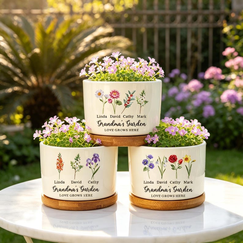 K&ouml;nnte beinhalten: Drei cremefarbene Keramik-Blument&ouml;pfe mit floralen Designs und dem Text "Grandma's Garden". Jeder Topf enth&auml;lt rosa bl&uuml;hende Pflanzen und steht auf einem runden Holzsockel. Der Text "Love Grows Here" ist ebenfalls aufgedruckt.
