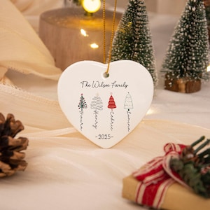 May include: A white heart-shaped ceramic ornament hanging by a gold string. It features the text The Wilson Family, the year 2025, and four illustrated Christmas trees with the names Maura, Henry, Cedric, and Lumina written vertically below.