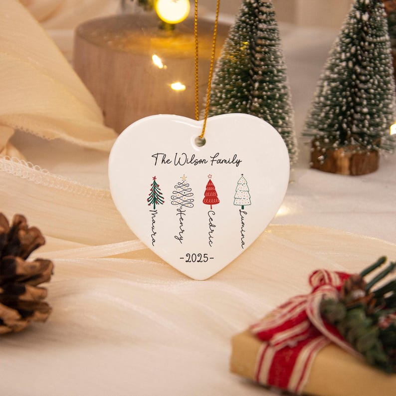 May include: A white heart-shaped ceramic ornament hanging by a gold string. It features the text The Wilson Family, the year 2025, and four illustrated Christmas trees with the names Maura, Henry, Cedric, and Lumina written vertically below.