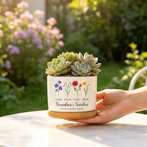 K&ouml;nnte beinhalten: Ein wei&szlig;er Keramik-Blumentopf mit Bambusfu&szlig;, gef&uuml;llt mit Sukkulenten. Der Topf ist mit Blumenillustrationen und dem Text "Grandma's Garden, Love Grows Here" mit Namen verziert. Der Topf wird von einer Person gehalten.