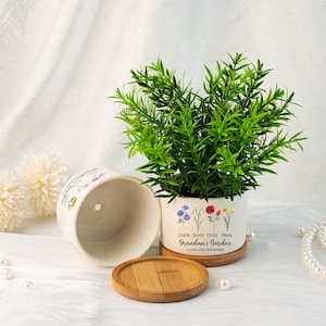 K&ouml;nnte beinhalten: Ein wei&szlig;er Keramik-Blumentopf mit einer gr&uuml;nen Pflanze, einem Untersetzer aus Holz und einem passenden leeren Topf. Der Topf ist mit floralen Illustrationen und dem Text "Grandma's Garden LOVE GROWS HERE" mit Namen verziert. Der Topf hat einen Durchmesser von ca. 13 cm.