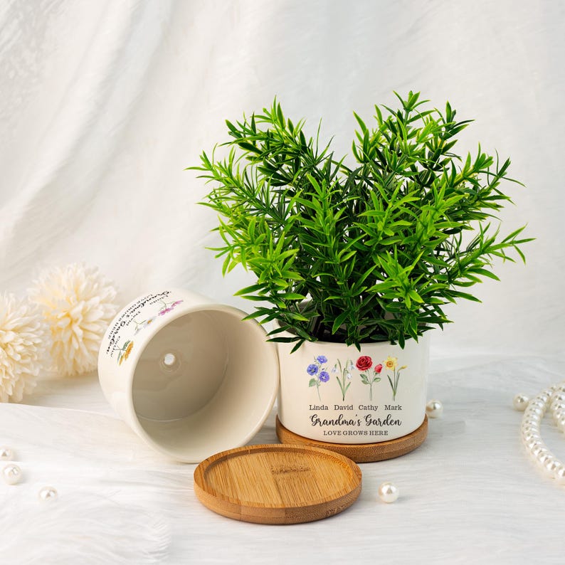 K&ouml;nnte beinhalten: Ein wei&szlig;er Keramik-Blumentopf mit einer gr&uuml;nen Pflanze, einem Untersetzer aus Holz und einem passenden leeren Topf. Der Topf ist mit floralen Illustrationen und dem Text "Grandma's Garden LOVE GROWS HERE" mit Namen verziert. Der Topf hat einen Durchmesser von ca. 13 cm.