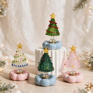 Op de afbeelding: Vier miniatuur kerstboomornamenten gemaakt van gewikkeld draad en glas. Elke boom heeft een gouden ster als piek en staat op een afgeronde, wolkachtige voet in pastelroze of lichtblauw. De bomen zijn uitgevoerd in groen, roze en helder glas.