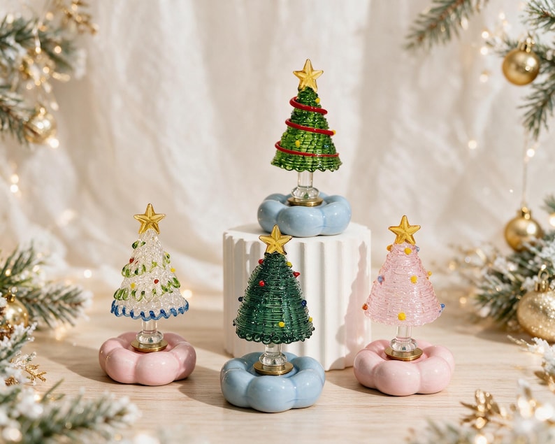 Op de afbeelding: Vier miniatuur kerstboomornamenten gemaakt van gewikkeld draad en glas. Elke boom heeft een gouden ster als piek en staat op een afgeronde, wolkachtige voet in pastelroze of lichtblauw. De bomen zijn uitgevoerd in groen, roze en helder glas.