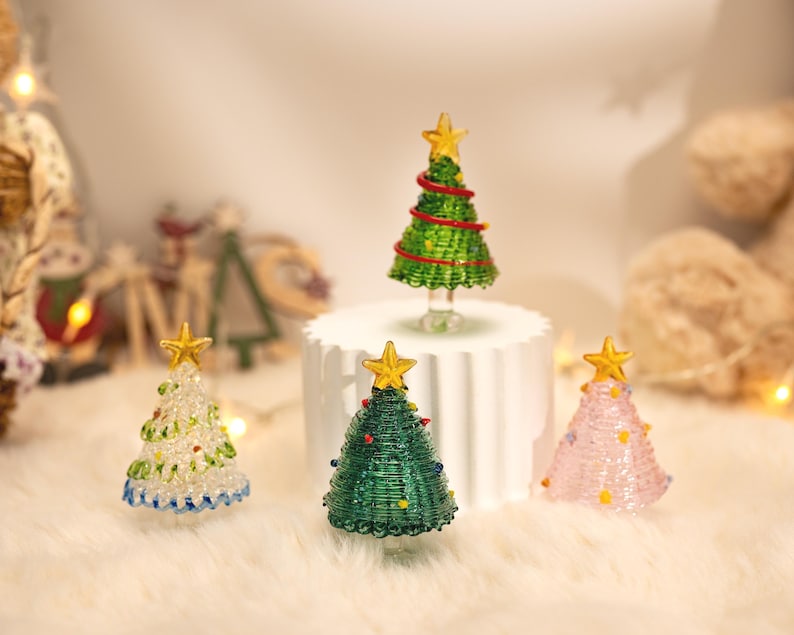 Op de afbeelding: Vier miniatuur kerstboombeeldjes gemaakt van gewikkeld glasdraad. E&eacute;n boom is felgroen met een rode spiraal, &eacute;&eacute;n is donkergroen met kleurrijke stippen, &eacute;&eacute;n is doorzichtig met groene accenten en &eacute;&eacute;n is lichtroze, allemaal bekroond met gouden sterren.