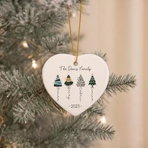 May include: A white heart-shaped ceramic ornament hanging by a gold string. It features the text The Davis Family, the year 2025, and four illustrated Christmas trees with the names Maura, Henry, Cedric, and Lumina written vertically below.