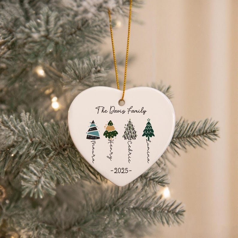 May include: A white heart-shaped ceramic ornament hanging by a gold string. It features the text The Davis Family, the year 2025, and four illustrated Christmas trees with the names Maura, Henry, Cedric, and Lumina written vertically below.