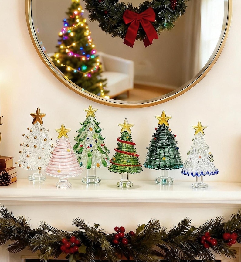 Op de afbeelding: Een collectie van zes decoratieve glazen kerstboombeeldjes, elk bekroond met een gouden ster. De bomen hebben diverse ontwerpen, waaronder helder glas met gouden stippen, roze horizontale strepen, groene takken en blauwe randaccenten.