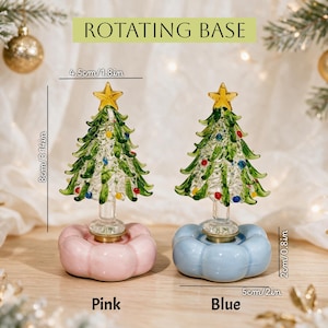 Op de afbeelding: Twee glazen kerstboomornamenten met gele sterren als piek, groene takken en kleurrijke accenten. Elke boom staat op een draaibare voet, &eacute;&eacute;n roze en &eacute;&eacute;n blauw. De bomen zijn 8 cm hoog en 4,5 cm breed, met voetstukken van 2 cm hoog en 5 cm breed.