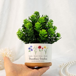 K&ouml;nnte beinhalten: Ein wei&szlig;er Keramik-Blumentopf mit Holzsockel, gef&uuml;llt mit leuchtend gr&uuml;nem Laub. Der Topf ist mit floralen Illustrationen und dem Text "Grandma's Garden, Love Grows Here" verziert. Namen sind &uuml;ber dem Text aufgedruckt. Der Topf wird in einer Hand gehalten.