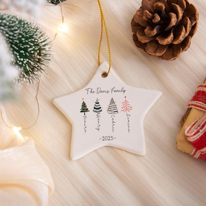 May include: A white star-shaped ceramic ornament hanging by a gold string. It features the text The Davis Family, the year 2025, and four illustrated Christmas trees with the names Maura, Henry, Cedric, and Lumina written vertically below.