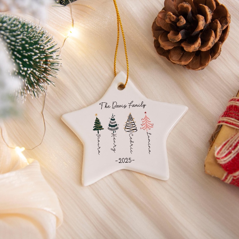 May include: A white star-shaped ceramic ornament hanging by a gold string. It features the text The Davis Family, the year 2025, and four illustrated Christmas trees with the names Maura, Henry, Cedric, and Lumina written vertically below.